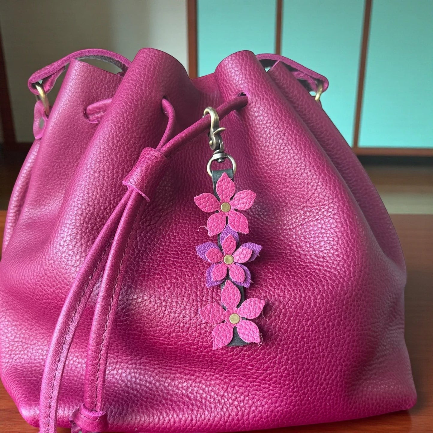Rhea Artistry Studio handmade leather floral charm with pink and purple flowers and a brushed brass clasp, attached to a magenta drawstring bag.