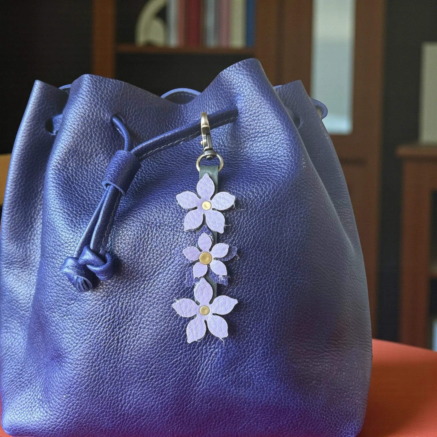 Rhea Artistry Studio handmade leather floral charm with three light purple flowers and brushed brass clasp, attached to a deep purple leather bucket bag.