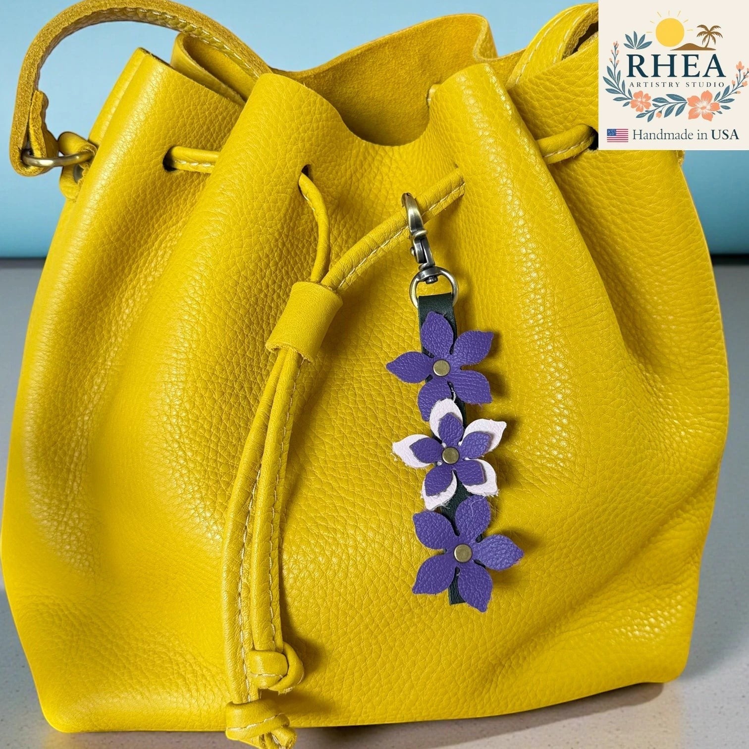 Rhea Artistry Studio handmade leather floral charm with purple and white flowers and a brushed brass clasp, attached to a yellow leather drawstring bag.