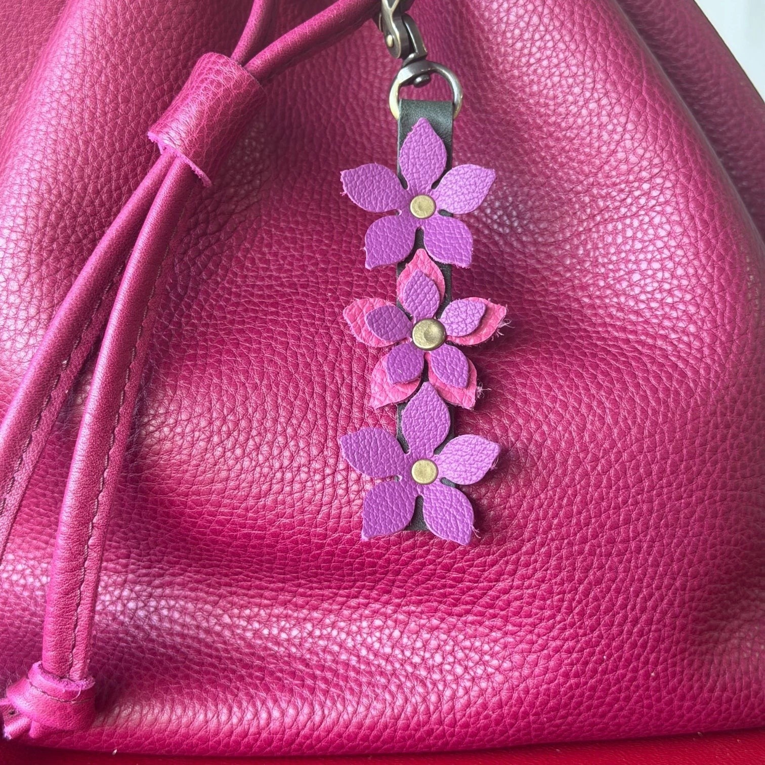 Rhea Artistry Studio handmade leather floral charm with purple and pink flowers and a brushed brass clasp, attached to a magenta leather bag.