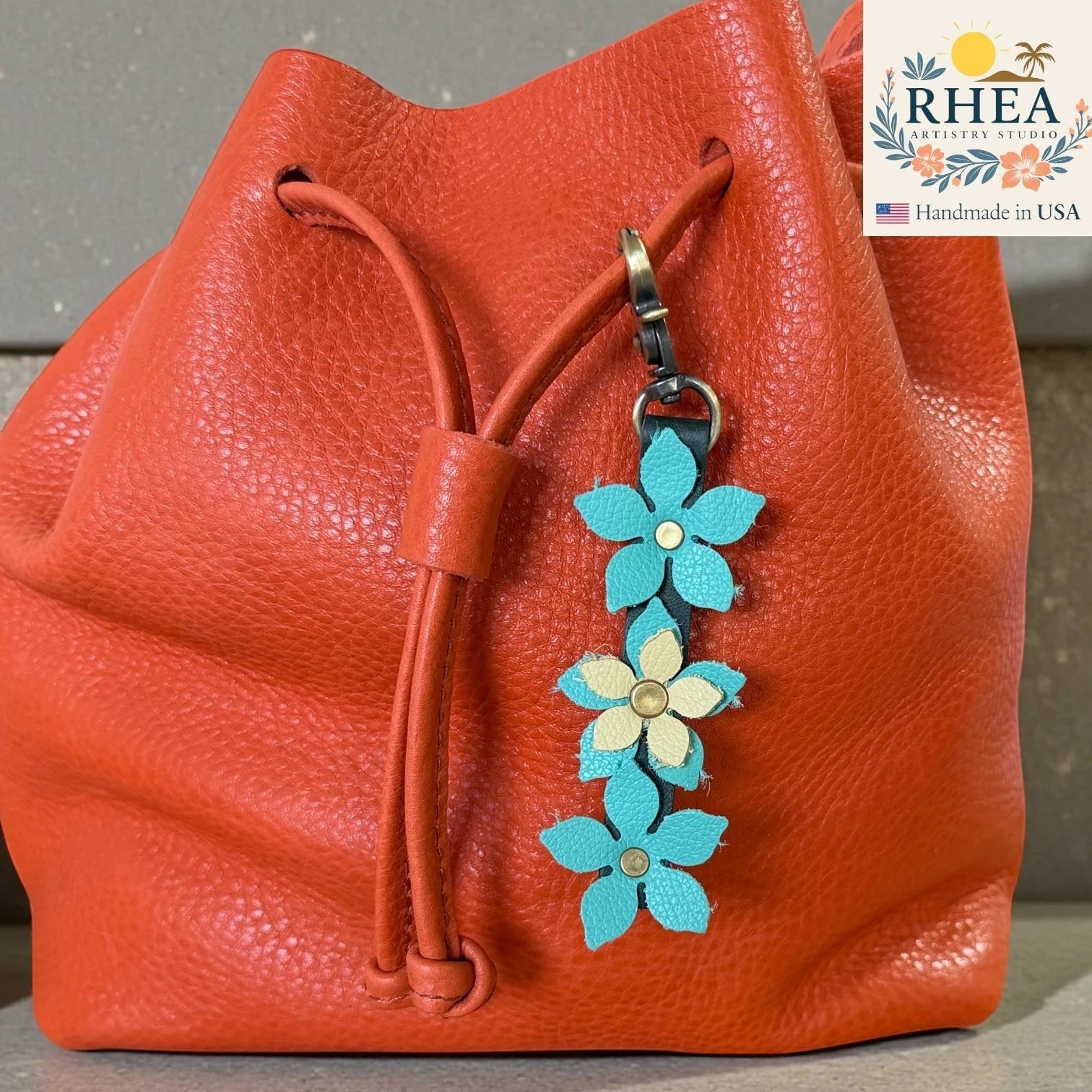Rhea Artistry Studio handmade leather floral charm with teal and cream flowers and a brushed brass clasp attached to an orange bucket bag.