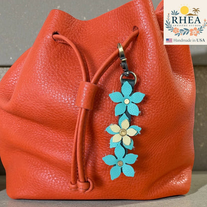 Rhea Artistry Studio handmade leather floral charm with teal and cream flowers and a brushed brass clasp attached to an orange bucket bag.