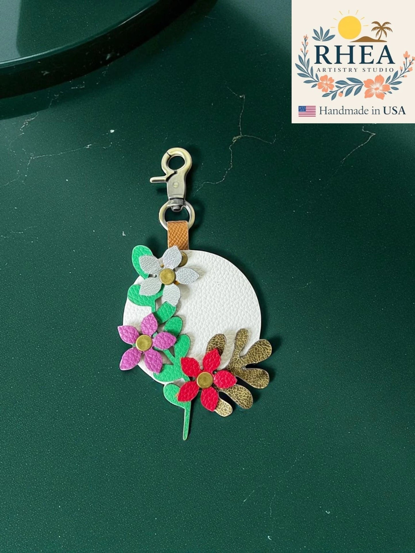 Rhea Artistry Studio white leather charm with layered floral appliques in pink, red, grey, green, and gold.