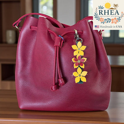 Rhea Artistry Studio handmade leather floral charm with yellow and red flowers and brushed brass clasp on a burgundy bucket bag.