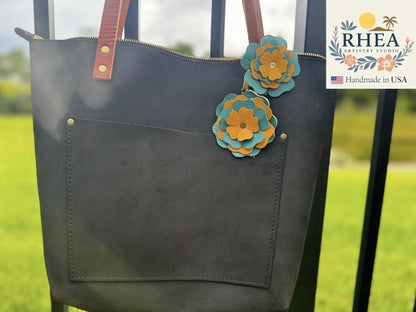 Rhea Artistry Studio handmade double leather flower bag charm in turquoise and yellow, attached to a black tote bag.