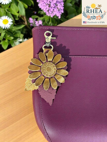 Rhea Artistry Studio daisy drop leather charm with gold petals and a textured purple leather leaf detail, attached to a plum handbag.