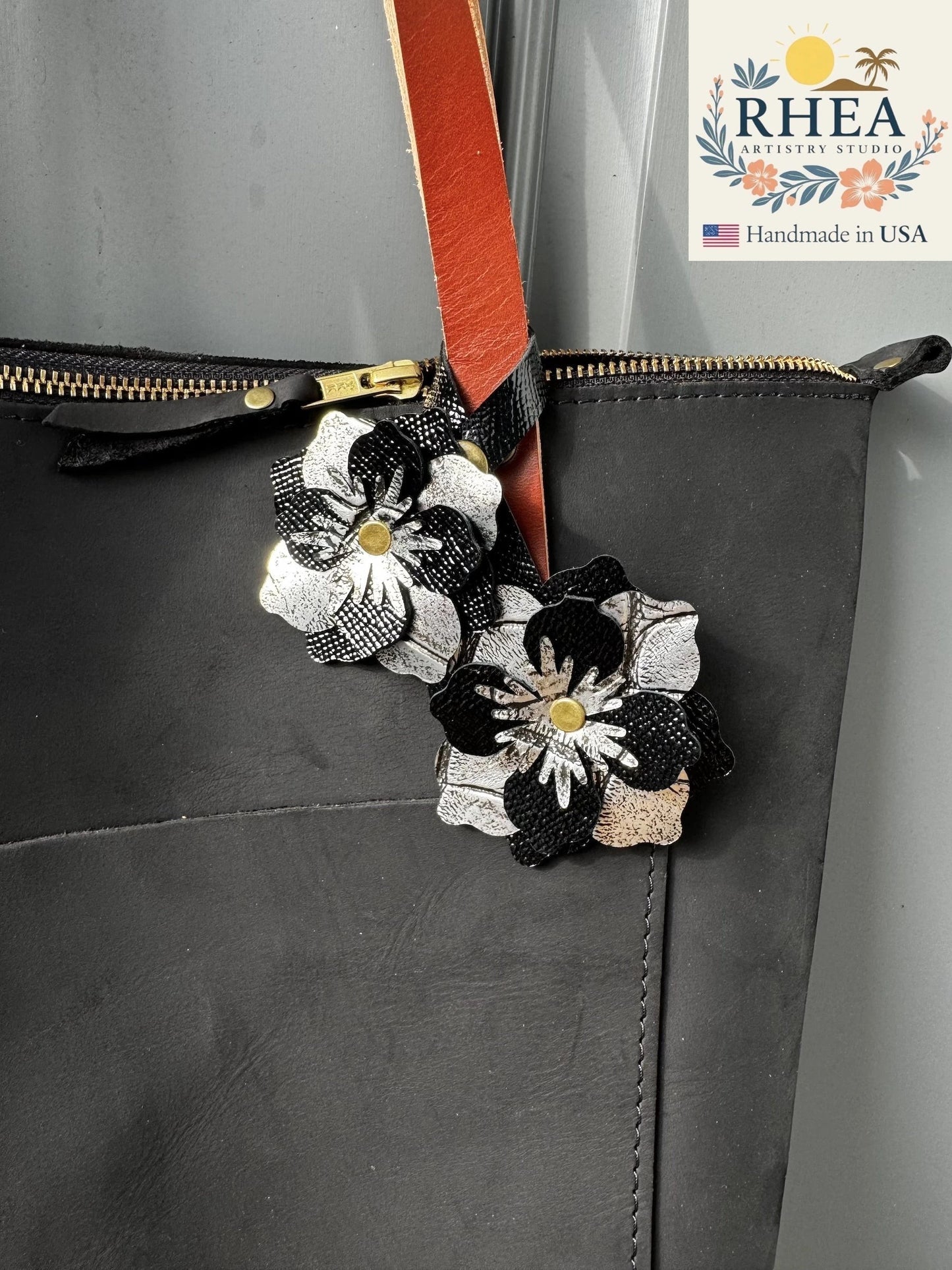 Rhea Artistry Studio black and silver leather flower double charm with brass rivets and snap fastener attached to a black tote bag.