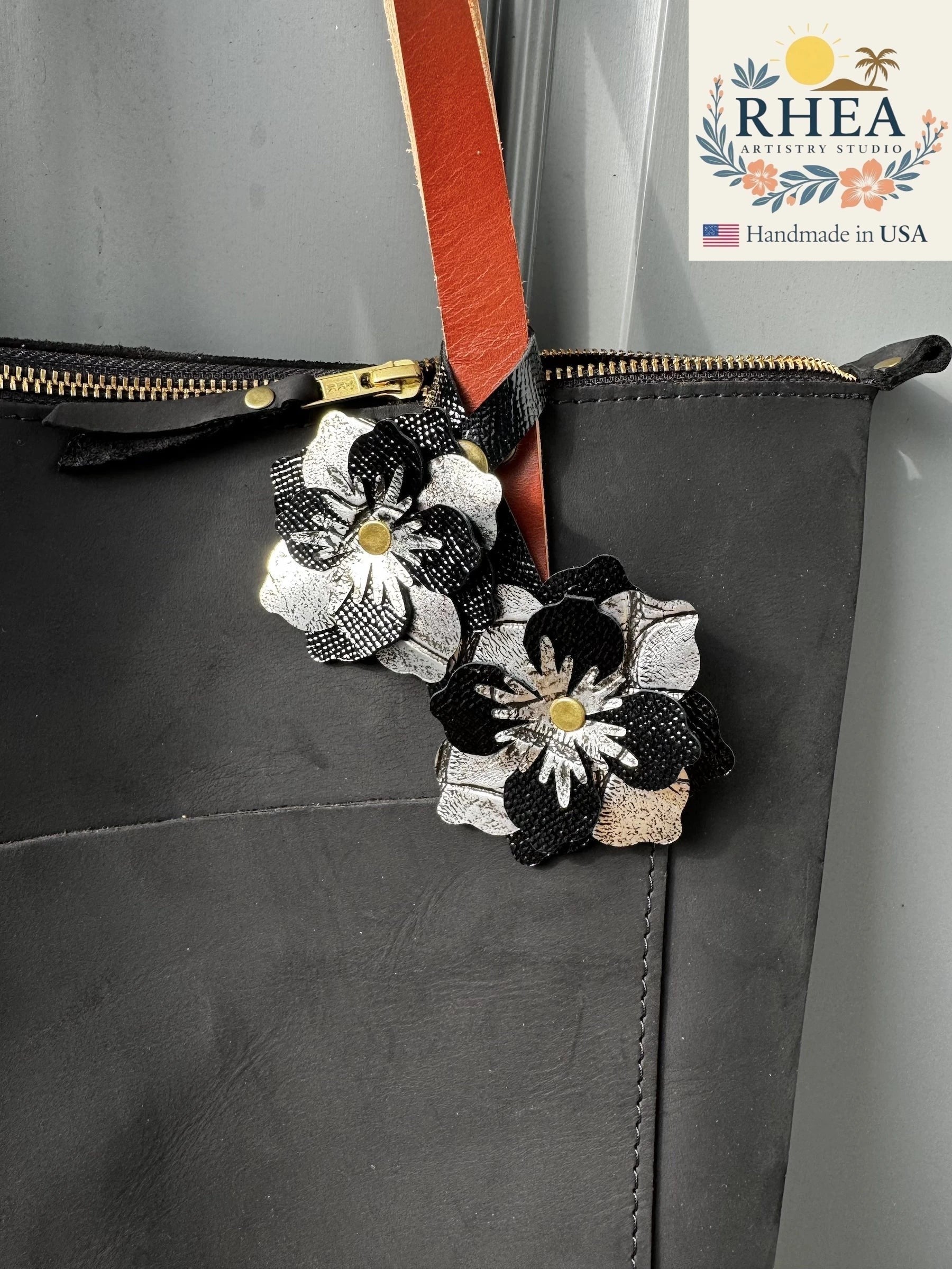 Rhea Artistry Studio black and silver leather flower double charm with brass rivets and snap fastener attached to a black tote bag.