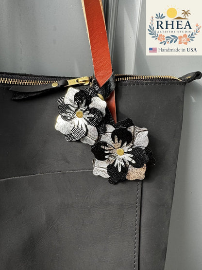 Rhea Artistry Studio black and silver leather flower double charm with brass rivets and snap fastener attached to a black tote bag.