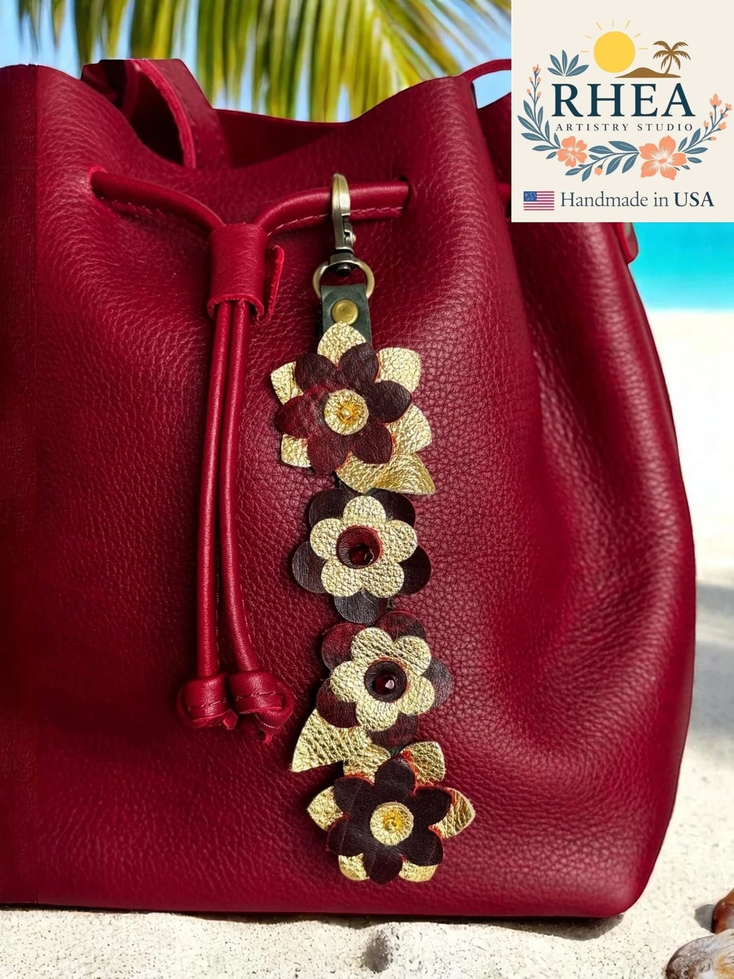Rhea Artistry Studio handcrafted leather flower charms in burgundy and gold metallic leather with Czech beads, attached to a red leather bucket bag.