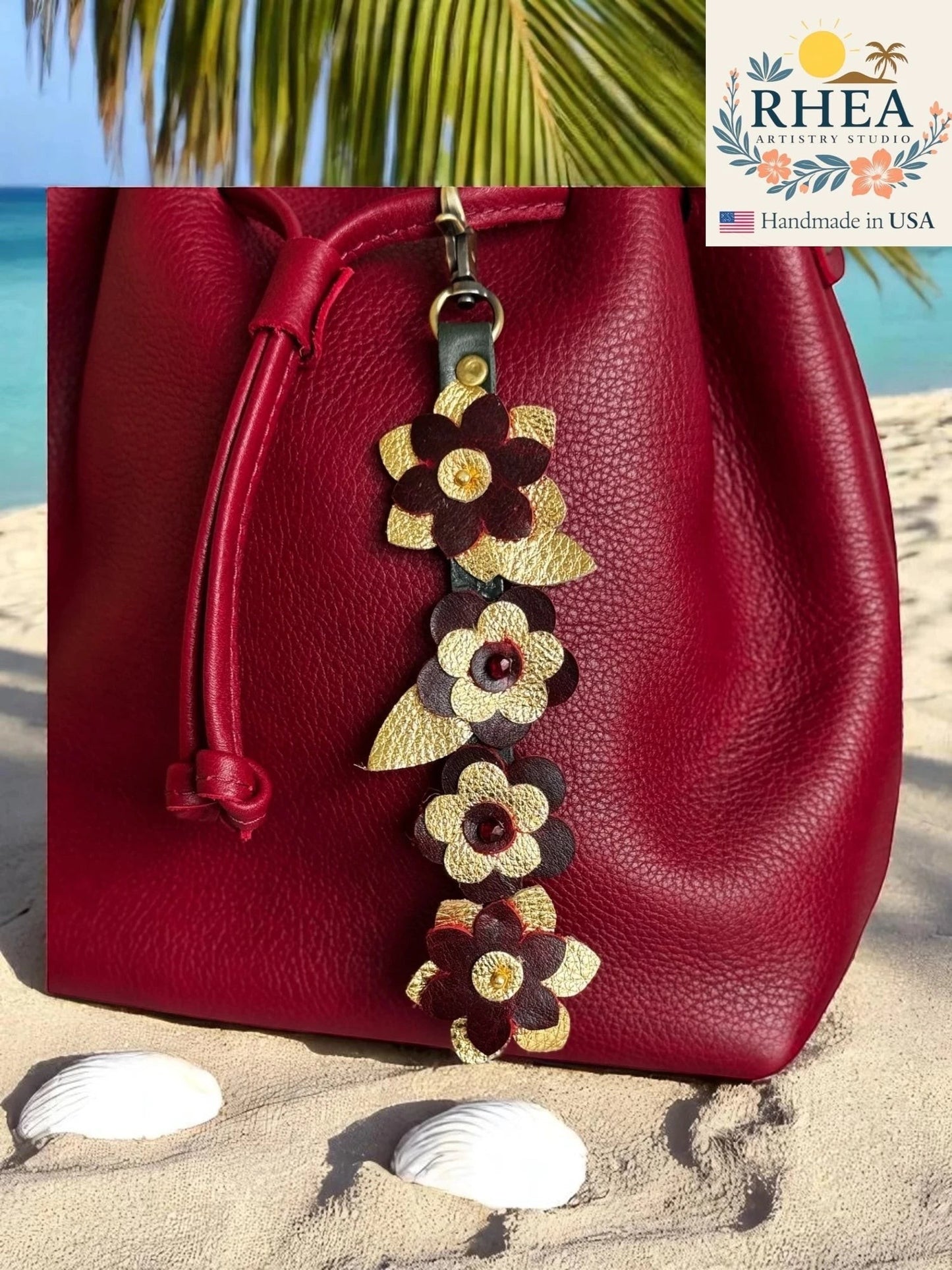 Rhea Artistry Studio handcrafted leather flower charm with Czech beads, featuring burgundy and gold petals, attached to a red leather bag on a sandy beach.