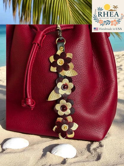 Rhea Artistry Studio handcrafted leather flower charm with Czech beads, featuring burgundy and gold petals, attached to a red leather bag on a sandy beach.