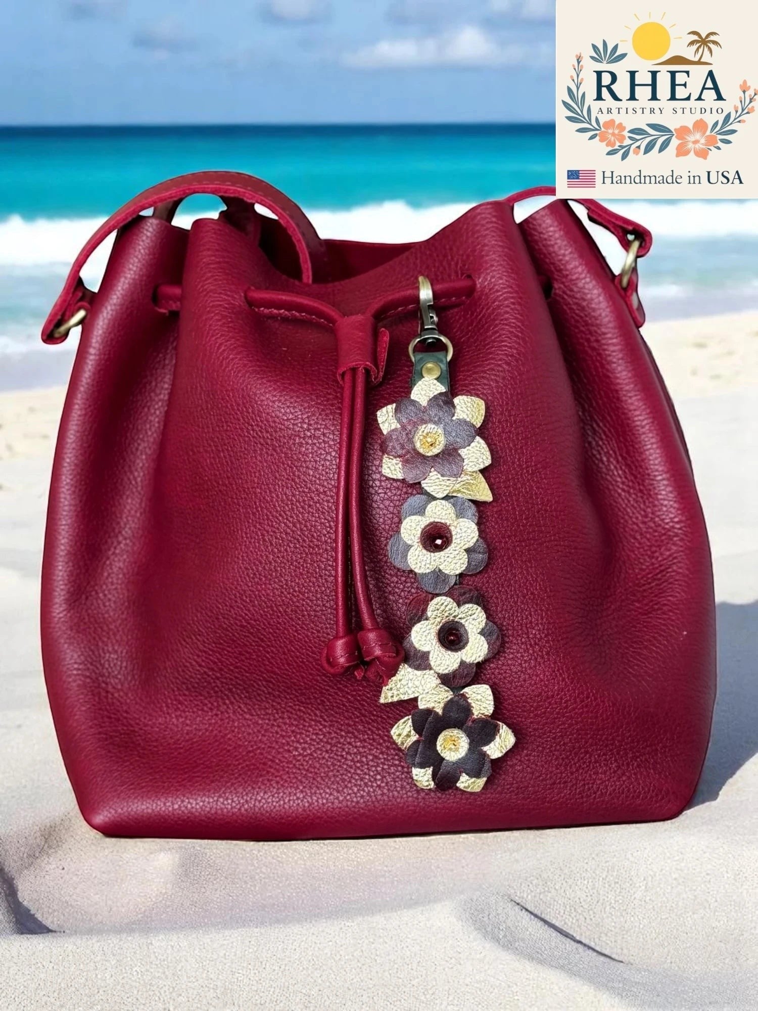 Rhea Artistry Studio handcrafted leather flower charms with Czech beads on a deep red leather bucket bag on a beach.