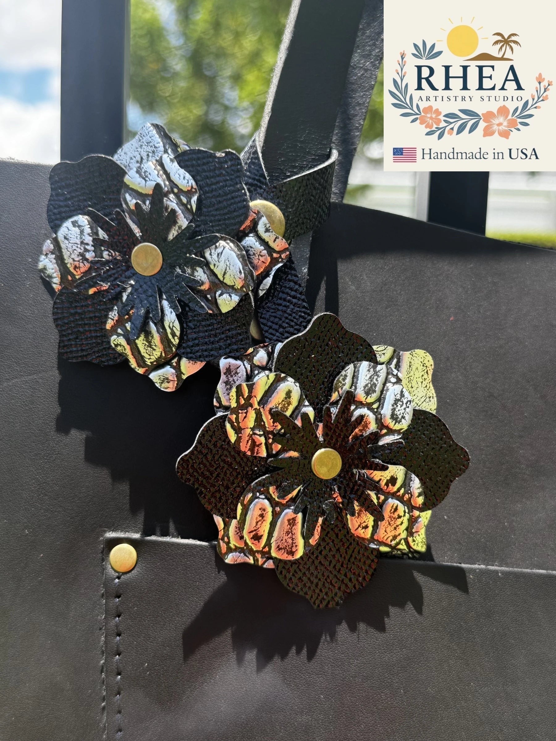Rhea Artistry Studio handcrafted double leather flower charm in silver and black with brass rivets, attached to a black leather bag.