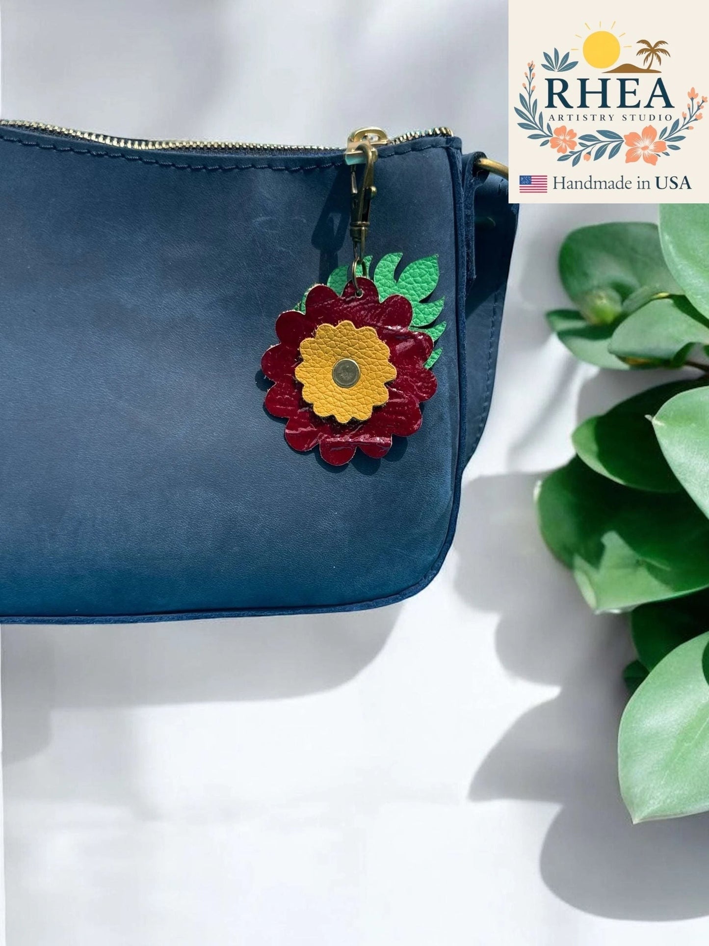 Rhea Artistry Studio leather monstera leaf and flower charm in burgundy and yellow with brass clasp, attached to a navy blue purse.