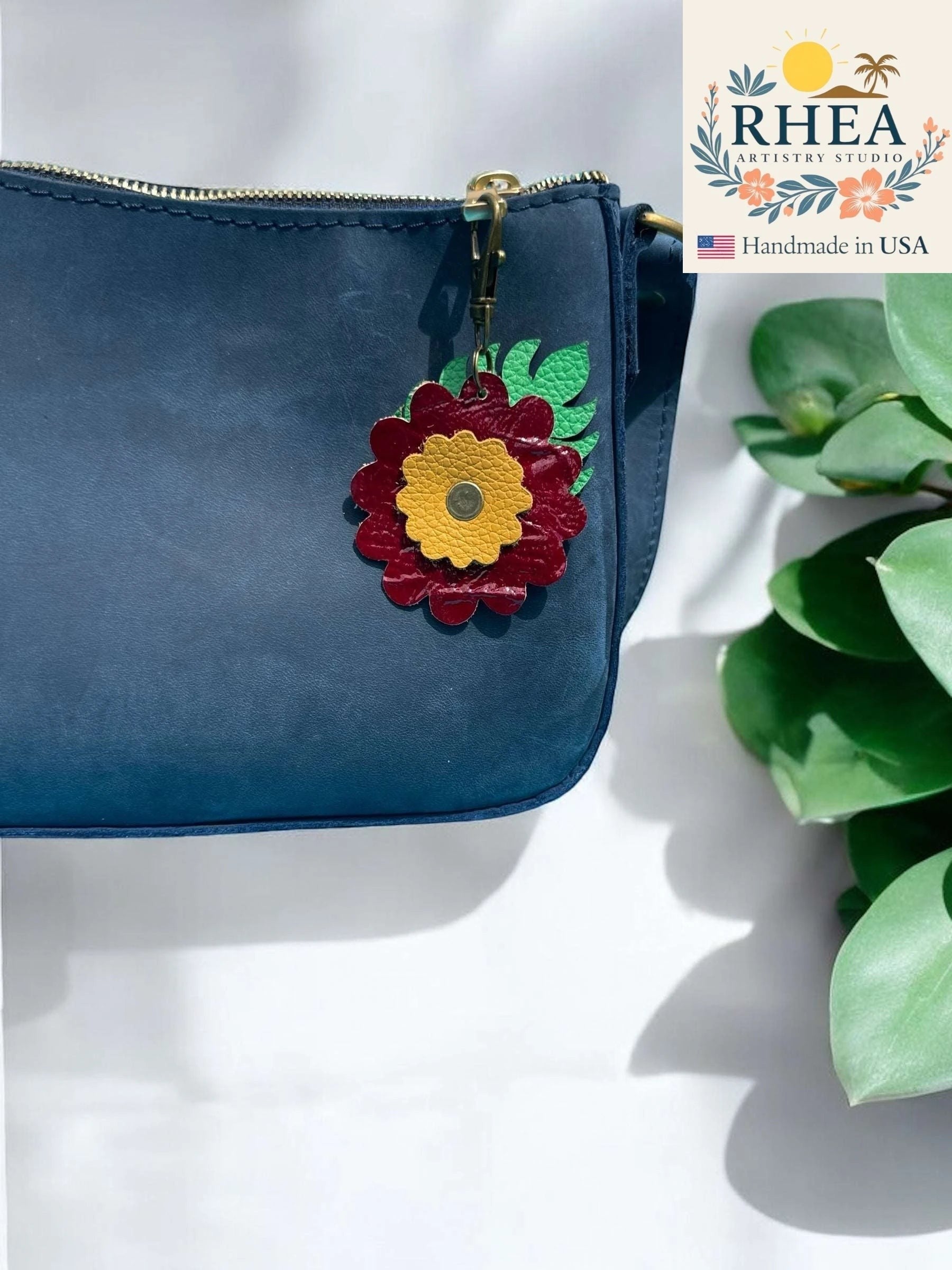 Rhea Artistry Studio leather monstera leaf and flower charm in burgundy and yellow with brass clasp, attached to a navy blue purse.
