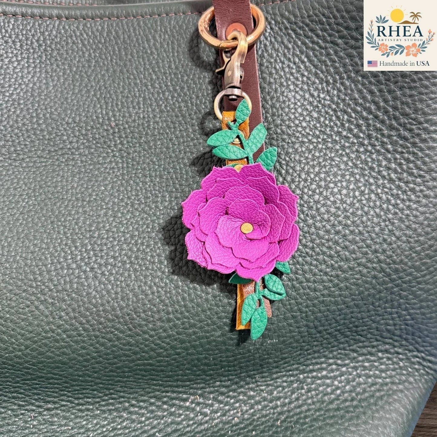 Rhea Artistry Studio handcrafted fuchsia leather flower keychain with green leaves and antique brass hardware, attached to a dark green pebbled leather tote bag.