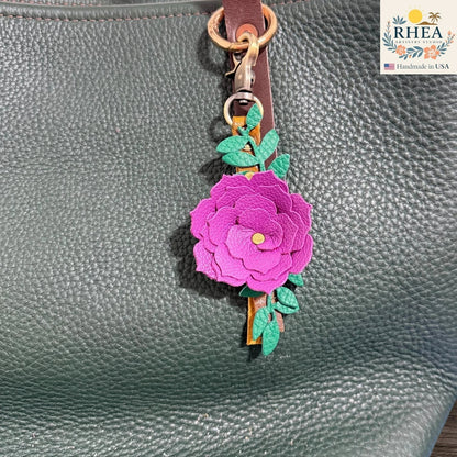 Rhea Artistry Studio handcrafted fuchsia leather flower keychain with green leaves and antique brass hardware, attached to a dark green pebbled leather tote bag.