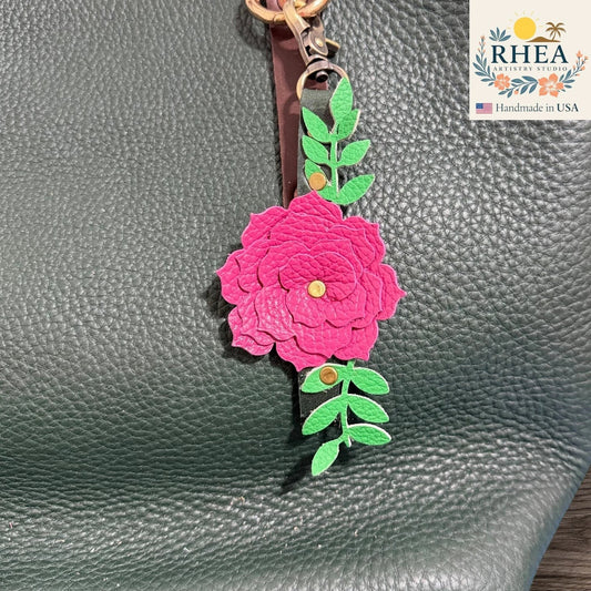 Rhea Artistry Studio handcrafted fuchsia leather flower keychain with green leather leaves and antique brass hardware attached to a dark green pebbled leather tote bag.