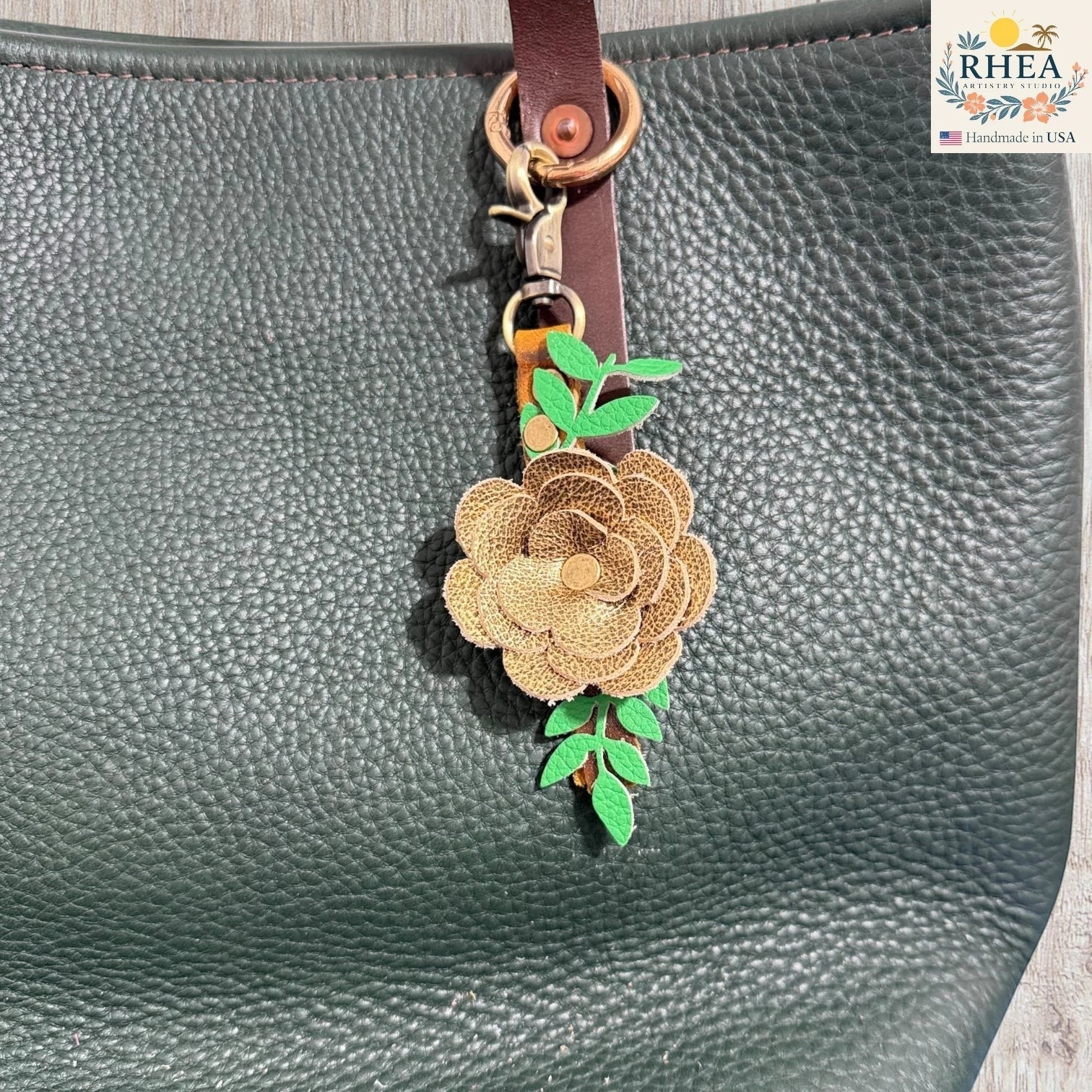 Rhea Artistry Studio handcrafted gold leather flower keychain with green leaves and antique brass hardware, attached to a dark green pebbled leather tote bag.