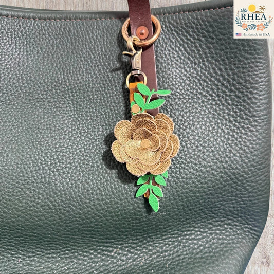 Rhea Artistry Studio handcrafted gold leather flower keychain with green leaves and antique brass hardware, attached to a dark green pebbled leather tote bag.