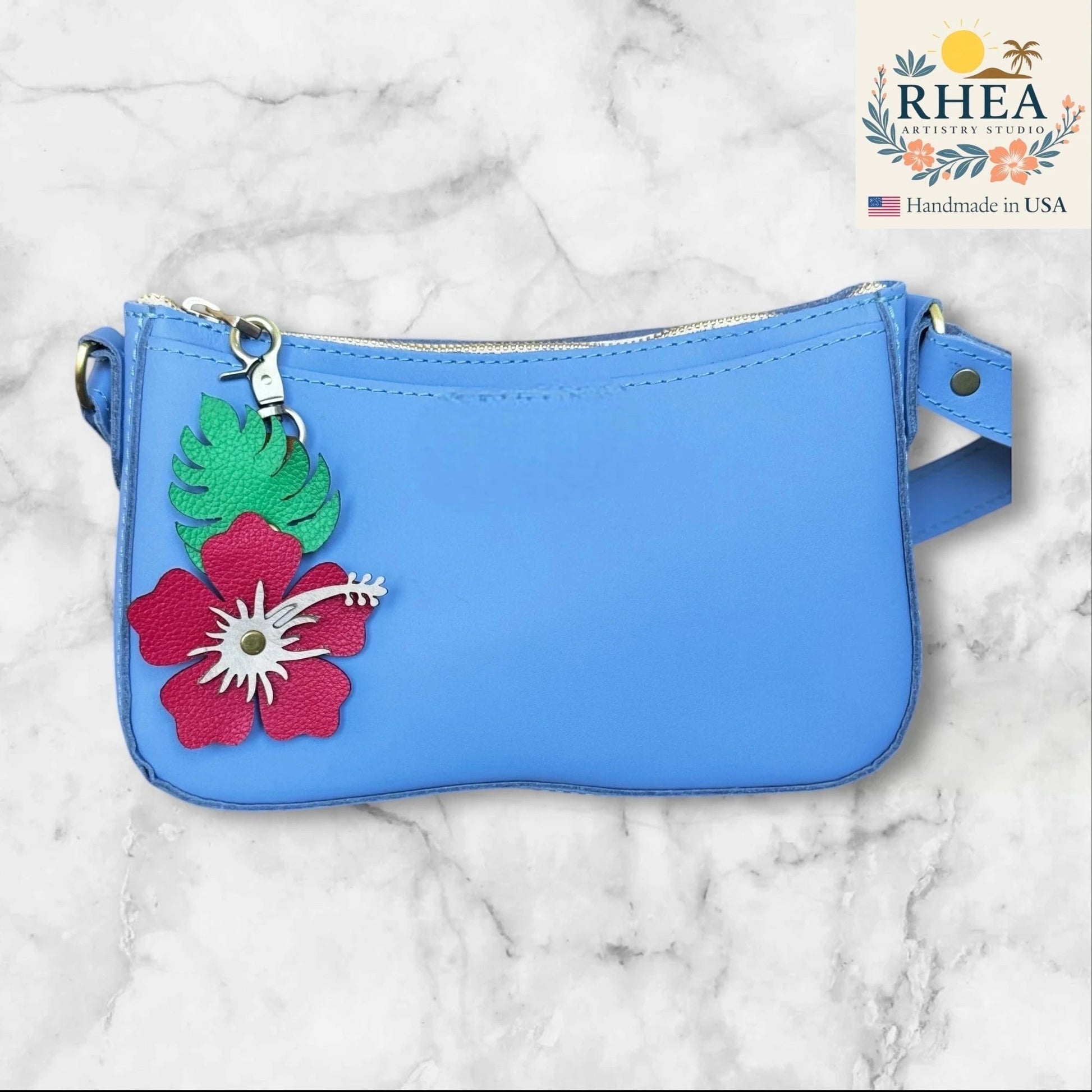 Rhea Artistry Studio blue leather crossbody bag with a handcrafted red hibiscus flower and green monstera leaf charm.