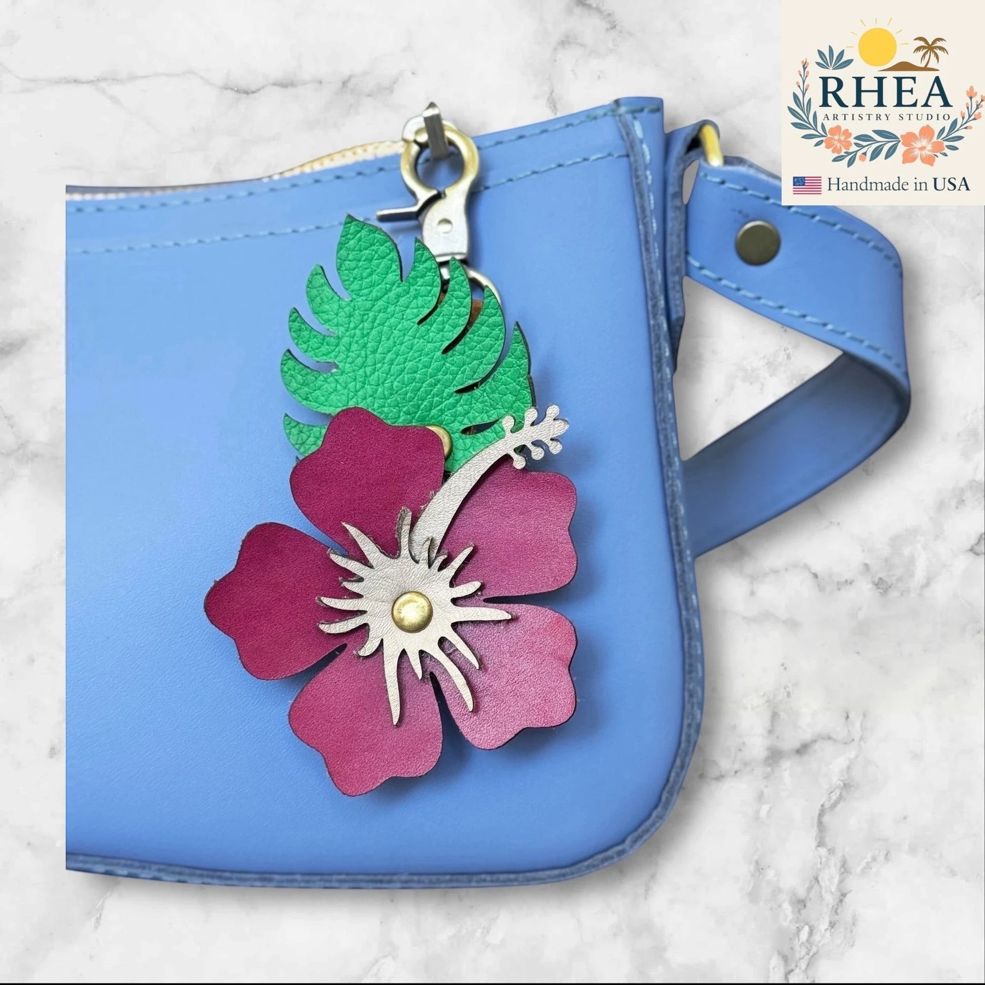 Rhea Artistry Studio handcrafted leather hibiscus charm with Monstera leaf, attached to a blue bag.