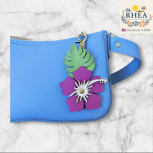 Rhea Artistry Studio handcrafted leather hibiscus charm with Monstera leaf on a blue bag. Features purple petals, silver stamen, and green leaf.