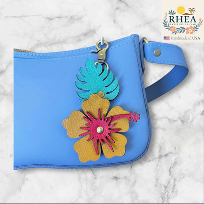 Rhea Artistry Studio handcrafted leather hibiscus charm with monstera leaf, yellow, pink, and teal colors, attached to a blue bag.