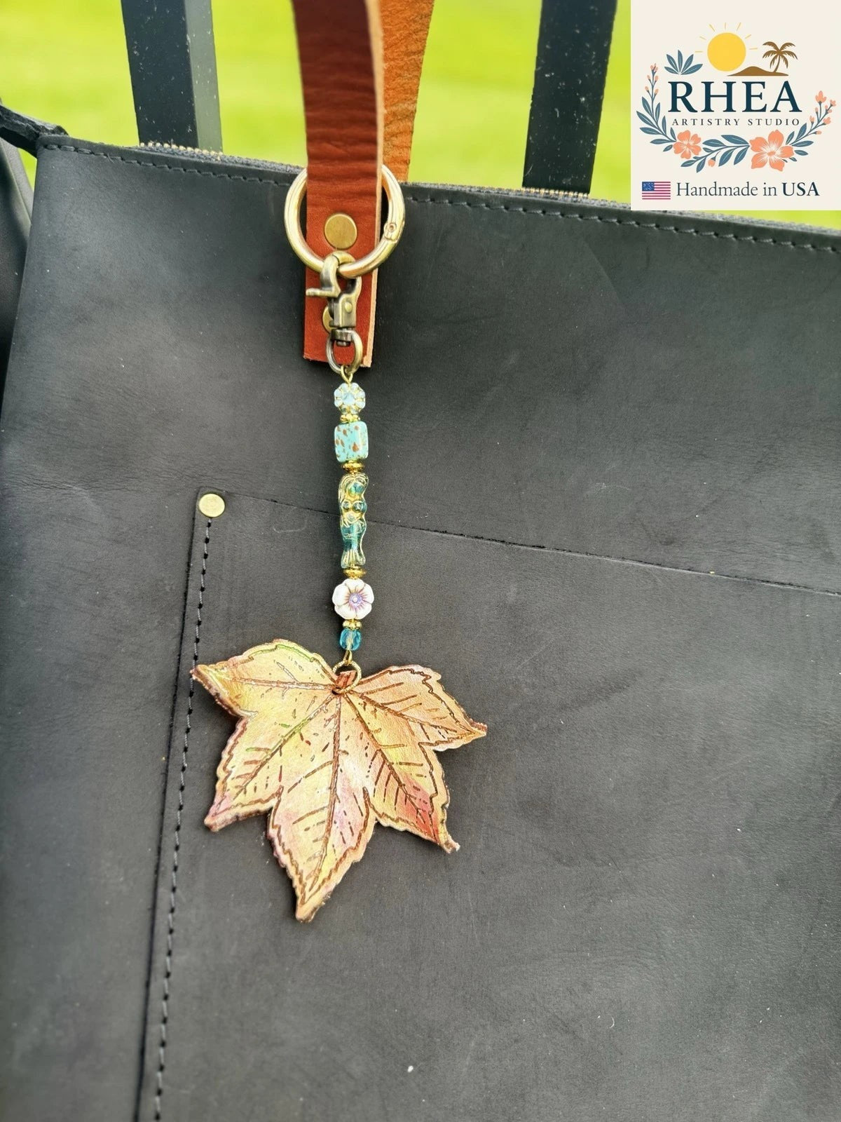 Rhea Artistry Studio Mermaid's Garden leather charm with maple leaf, Czech glass beads, and brass hardware on a black bag.