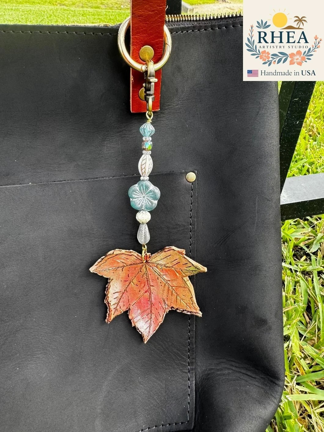 Rhea Artistry Studio Ocean Breeze Flora leather charm with a red and orange leaf pendant and blue glass beads on a black bag.