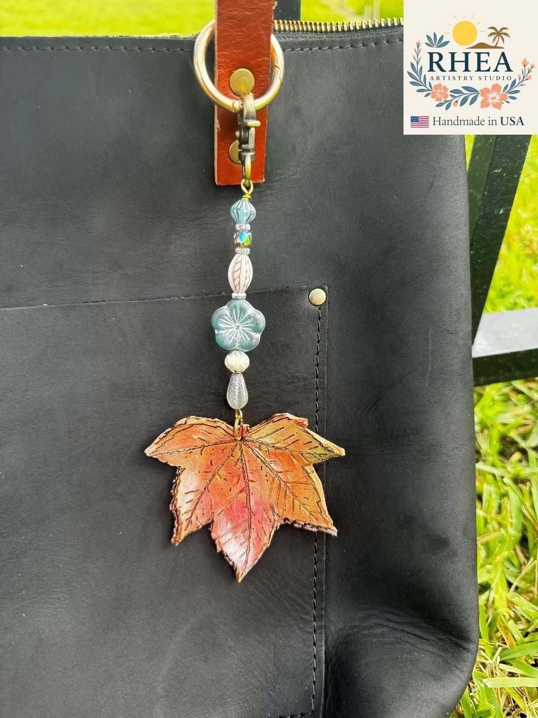 Rhea Artistry Studio Ocean Breeze Flora leather charm with a hand-painted maple leaf, blue floral bead, and brass hardware. Attached to a black leather bag.