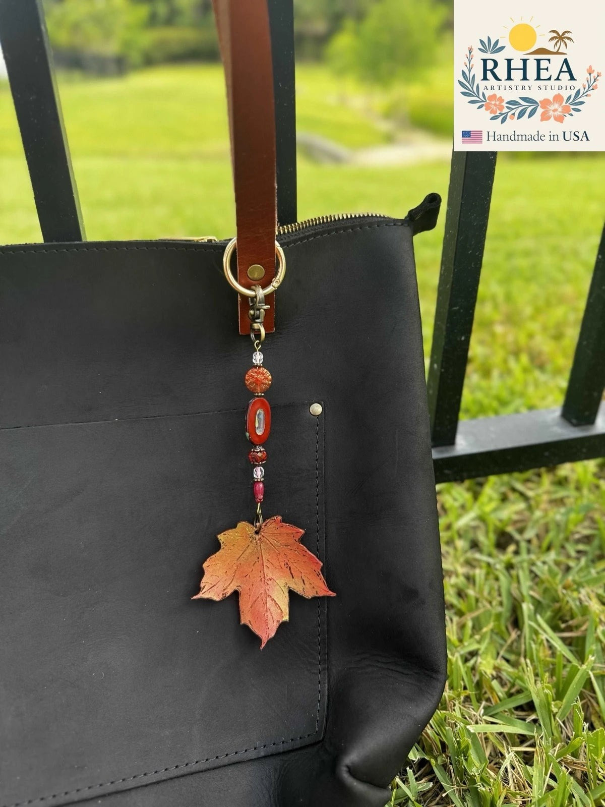 Rhea Artistry Studio Crimson Dahlia Delight leather charm with red and orange leaf, Czech glass beads, and brass hardware attached to a black bag.