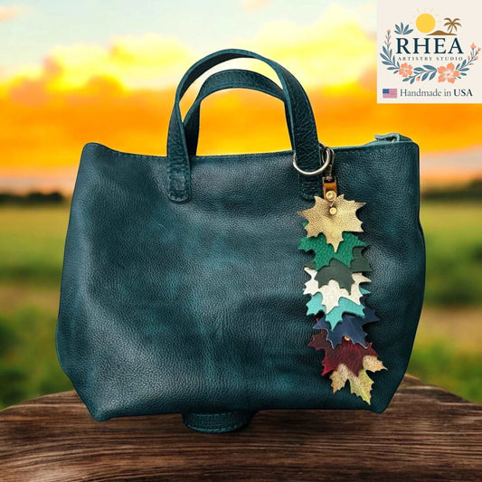 Rhea Artistry Studio handcrafted leather maple leaf bag charm in multi-color, attached to a teal tote bag.