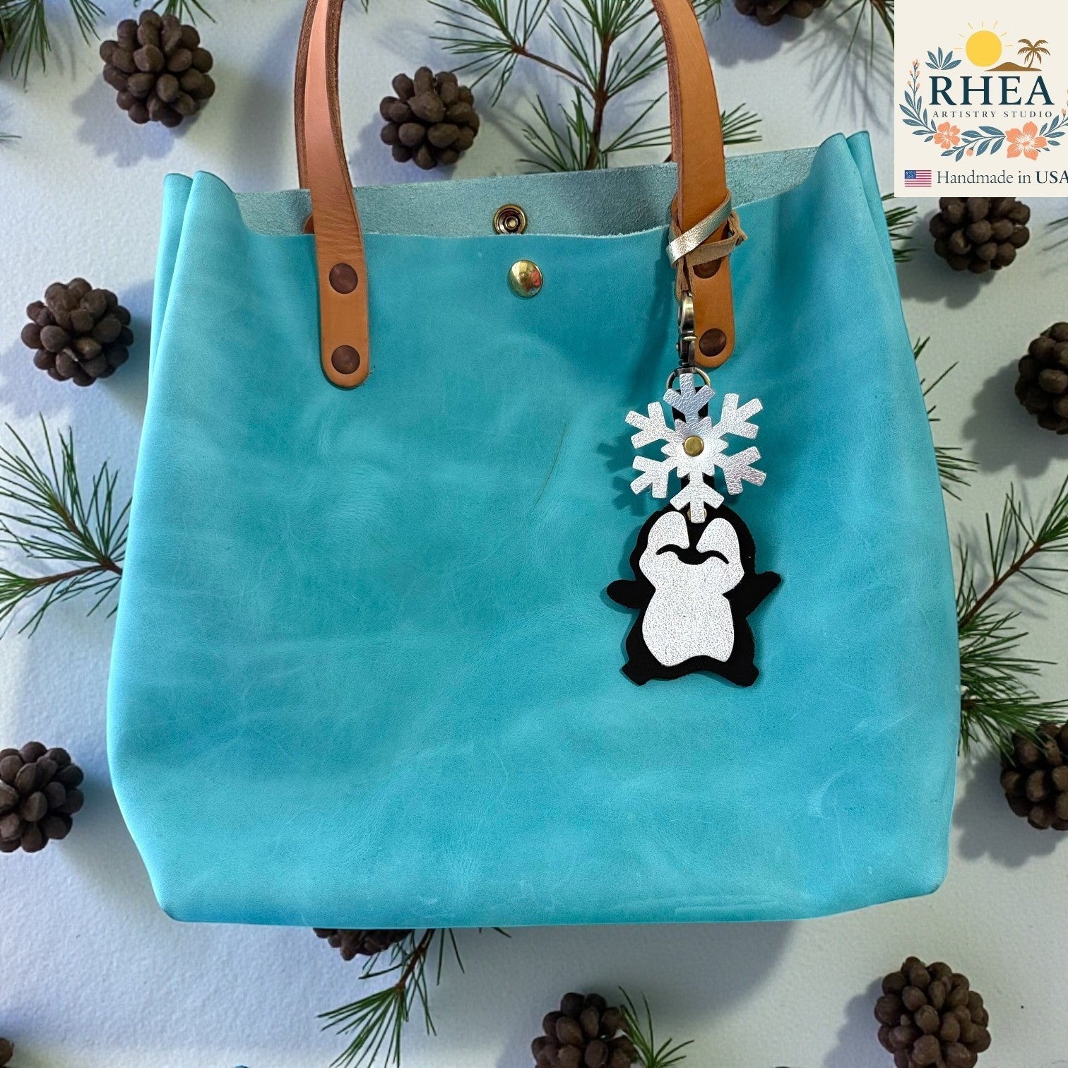 Rhea Artistry Studio handmade leather penguin charm with a silver glitter snowflake detail, attached to a turquoise tote bag.