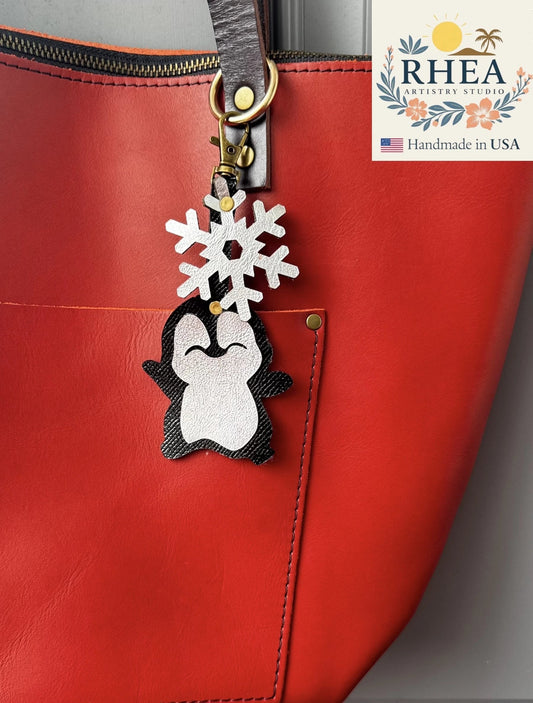 Rhea Artistry Studio handmade black and white leather penguin charm with a silver snowflake detail attached to a red tote bag.