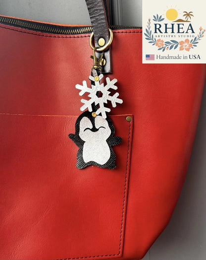 Rhea Artistry Studio handmade leather penguin charm with snowflake detail, attached to a red tote bag.