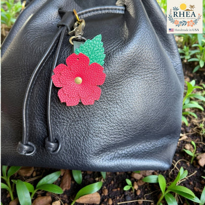 Rhea Artistry Studio red layered leather rose charm with green engraved leaf detail and brass hardware, attached to a black leather bag.
