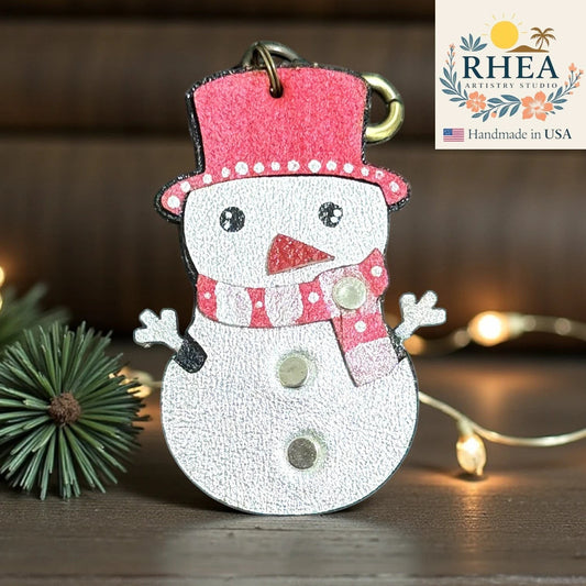 Rhea Artistry Studio handmade leather snowman charm with red hat and scarf, silver body, and button details. Festive winter accessory.