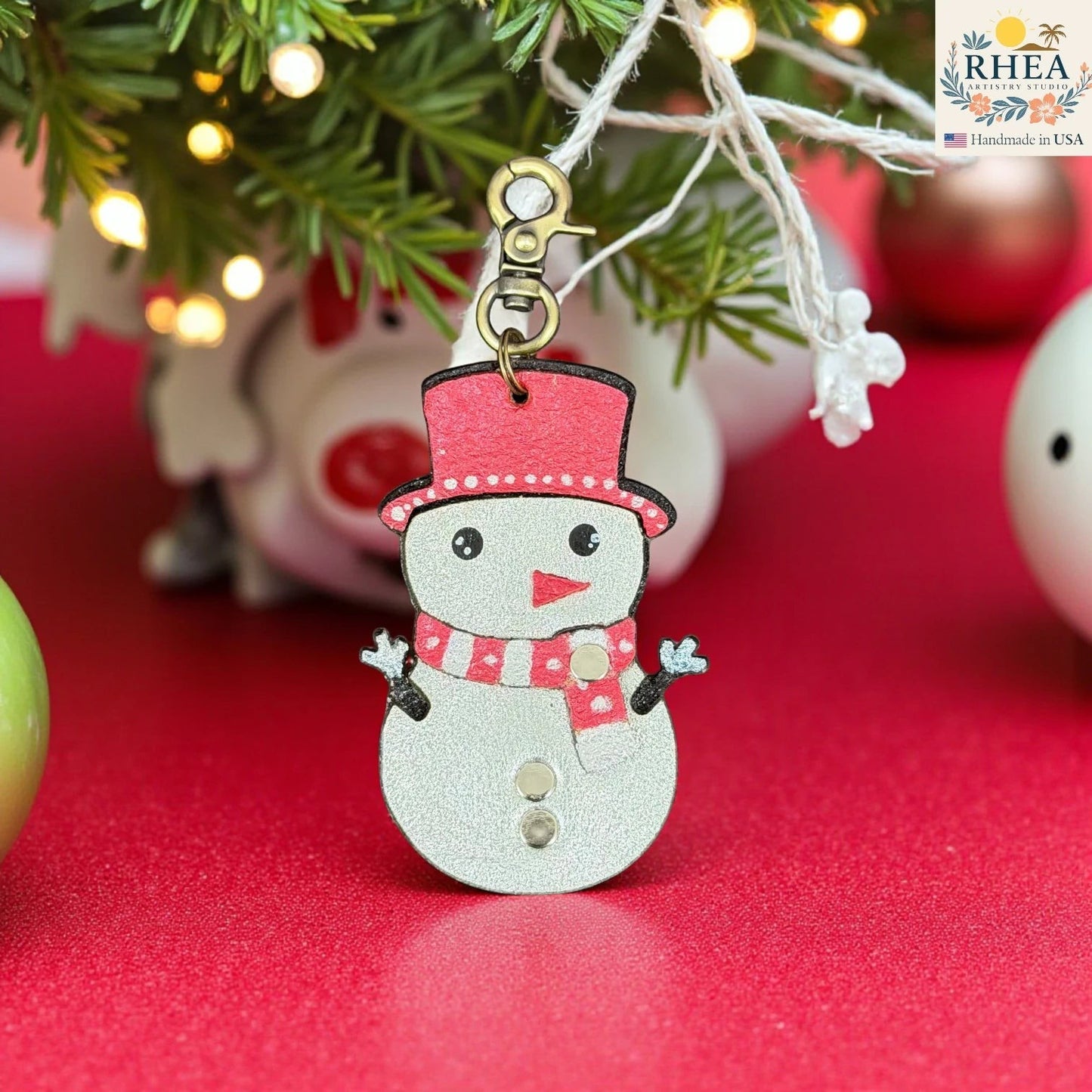 Rhea Artistry Studio handmade leather snowman charm with red hat and scarf, silver body, and rivet buttons. Festive winter accessory.