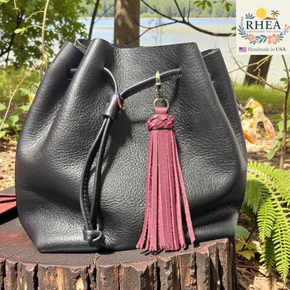 Rhea Artistry Studio burgundy leather tassel bag charm with braided detail and brass clasp, attached to a black leather drawstring bag.