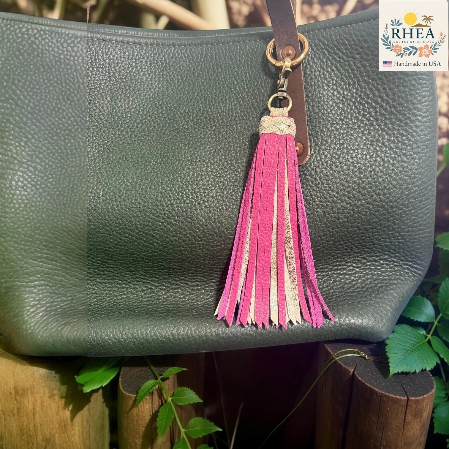 Rhea Artistry Studio handmade leather tassel charm with pink and metallic gold accents on a green pebbled leather bag.