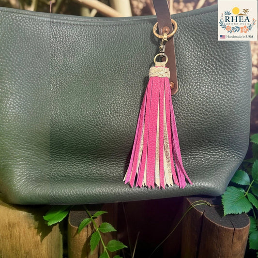 Rhea Artistry Studio handmade leather tassel charm with pink and metallic gold accents on a green pebbled leather bag.