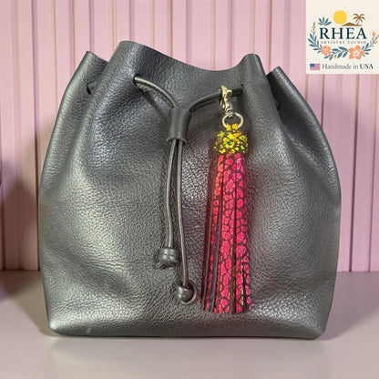 Rhea Artistry Studio handmade dual-tone leather tassel keychain with glitter finish and antique brass lobster clasp attached to a grey leather drawstring bag.