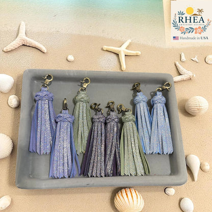 Rhea Artistry Studio handmade leather tassel keychains in pastel blue, sage green, and lavender with glitter accents and antique brass clasps.