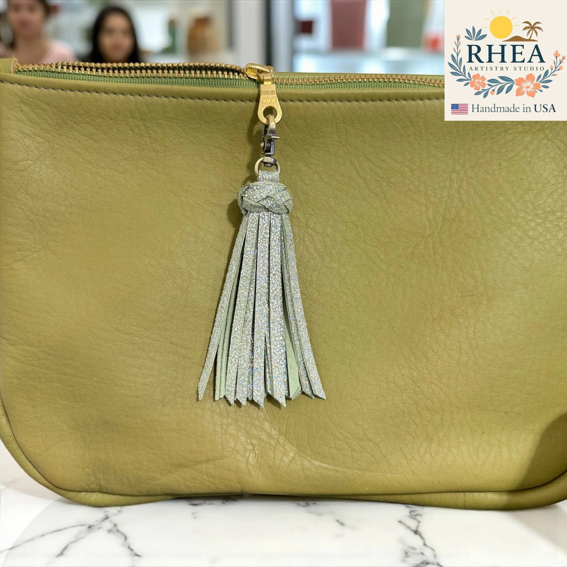 Rhea Artistry Studio handmade light green leather tassel keychain with glitter accents and antique brass lobster clasp attached to a light green leather bag.