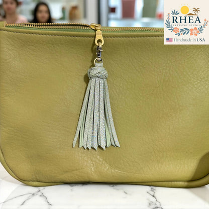Rhea Artistry Studio handmade light green leather tassel keychain with glitter accents and antique brass lobster clasp attached to a light green leather bag.