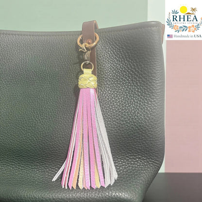 Rhea Artistry Studio handmade leather tassel keychain in pink, gold, and grey with a lobster clasp, attached to a black pebbled leather bag.