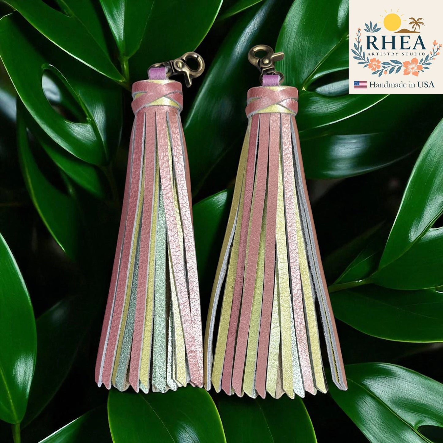 Rhea Artistry Studio handmade pink, yellow, and silver leather tassel keychain with lobster clasp on green leaves.