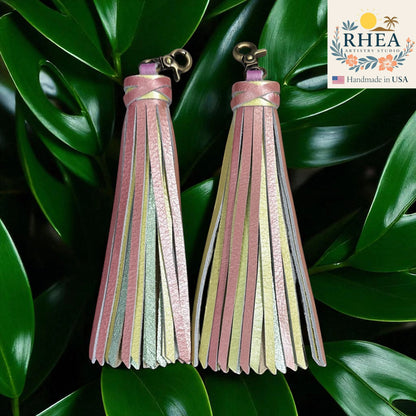 Rhea Artistry Studio handmade pink, yellow, and silver leather tassel keychain with lobster clasp on green leaves.